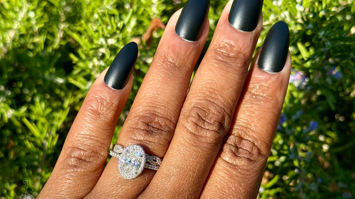 Hand with matte blackalmond-shaped nails, wearing a diamond ring, against a lush green background.