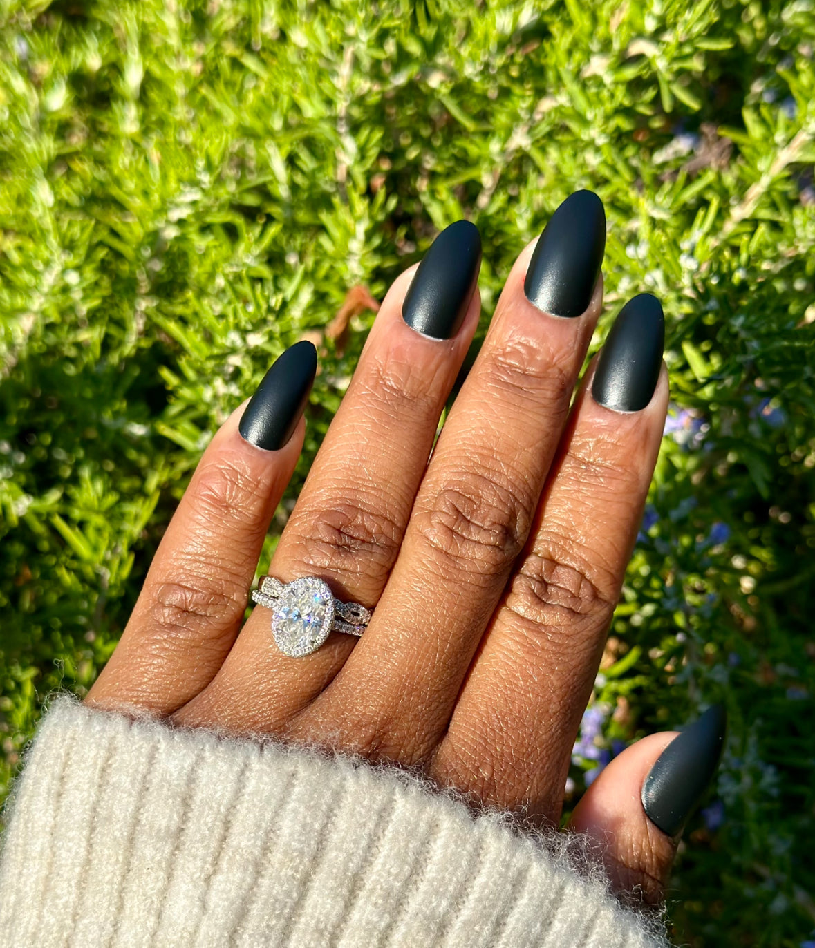Hand with matte blackalmond-shaped nails, wearing a diamond ring, against a lush green background.