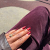 A close-up of a hand with glossy cat-eye dark red manicured nails resting on a person's lap wearing a deep plum corduroy pants, with a gold ring on the ring finger.