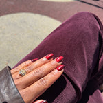 A close-up of a hand with glossy cat-eye dark red manicured nails resting on a person's lap wearing a deep plum corduroy pants, with a gold ring on the ring finger.
