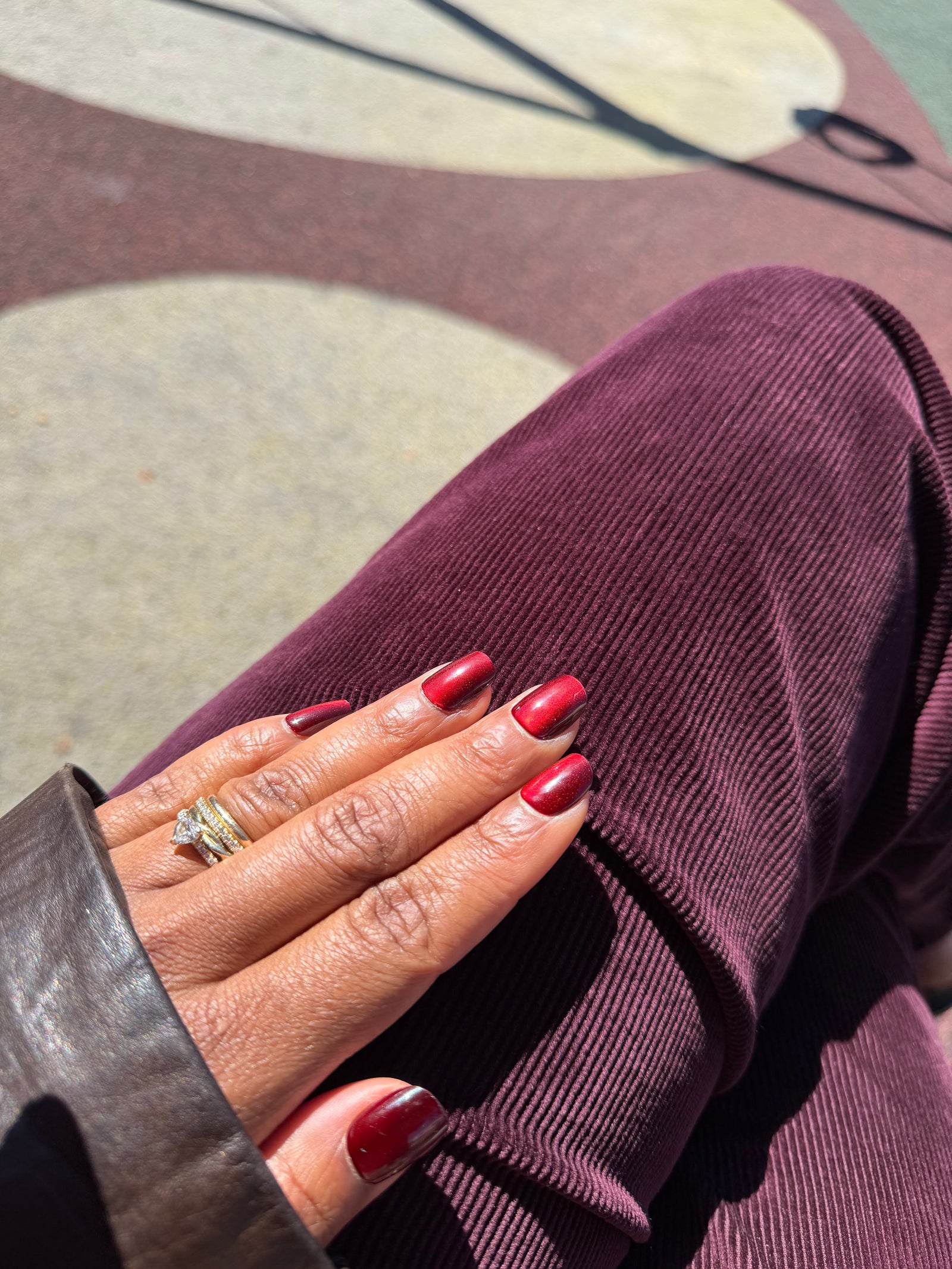 A close-up of a hand with glossy cat-eye dark red manicured nails resting on a person's lap wearing a deep plum corduroy pants, with a gold ring on the ring finger.