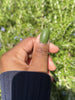 Thumb with olive green press-on nail, outdoor setting.